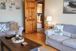 Una sala de estar acogedora con una puerta de madera, un sofá con un cojín que dice 'let's cuddle', una mesita con una lámpara y un tablero de ajedrez sobre una mesa de centro.