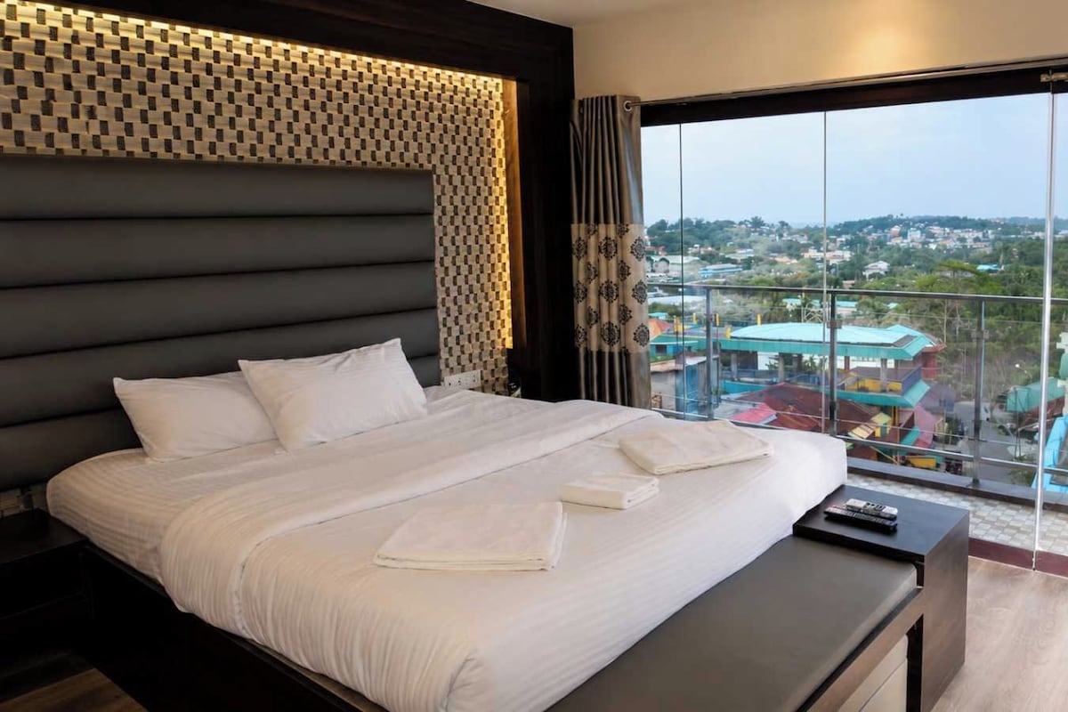 A hotel room with a large bed, a view of a cityscape through floor-to-ceiling windows, and a balcony with a railing.