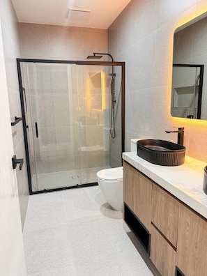 A modern bathroom with a glass-enclosed shower, a white toilet, and a wooden vanity with a black sink.