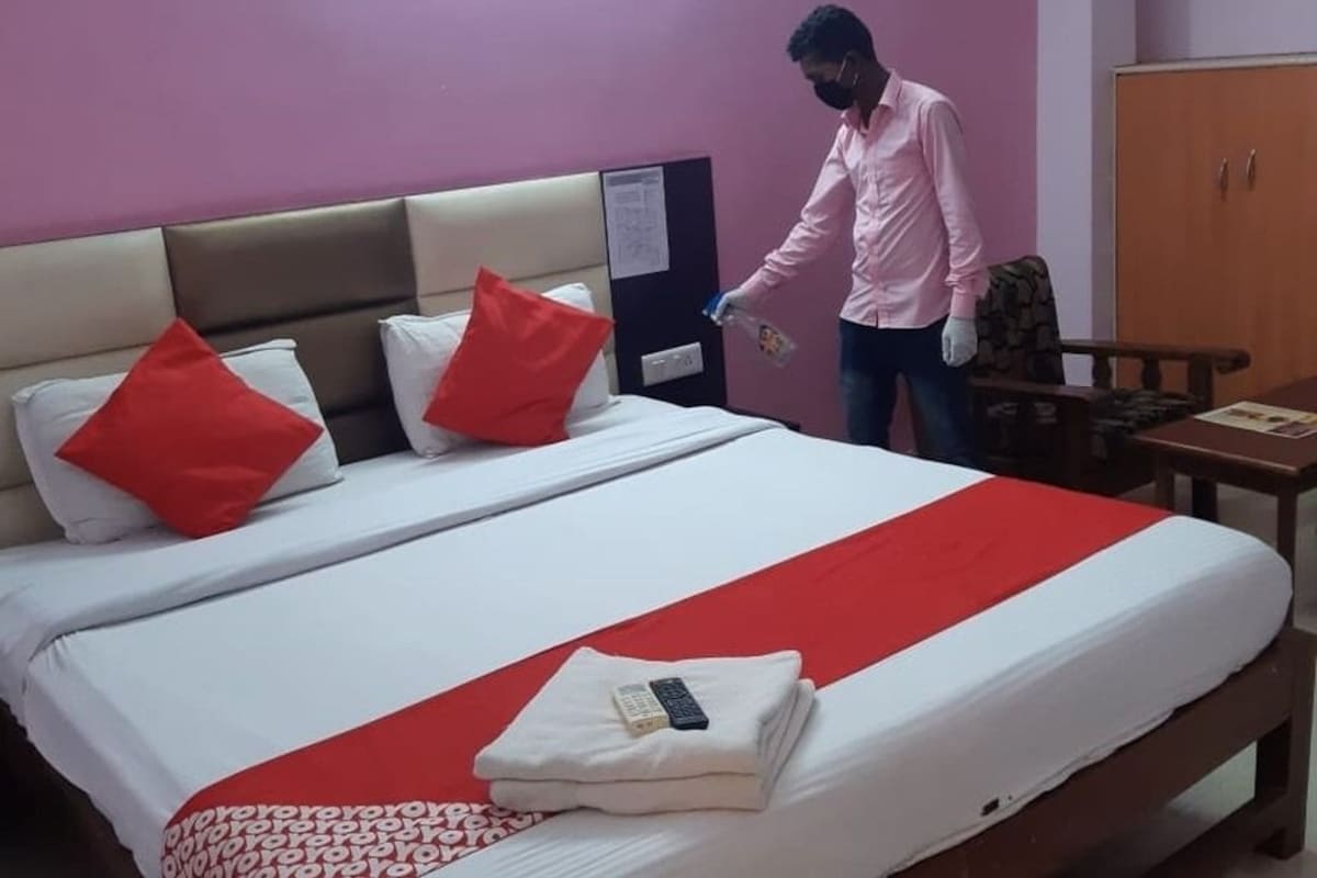 Quarto de hotel com cama, almofadas, manta vermelha, controle remoto e uma pessoa limpando.