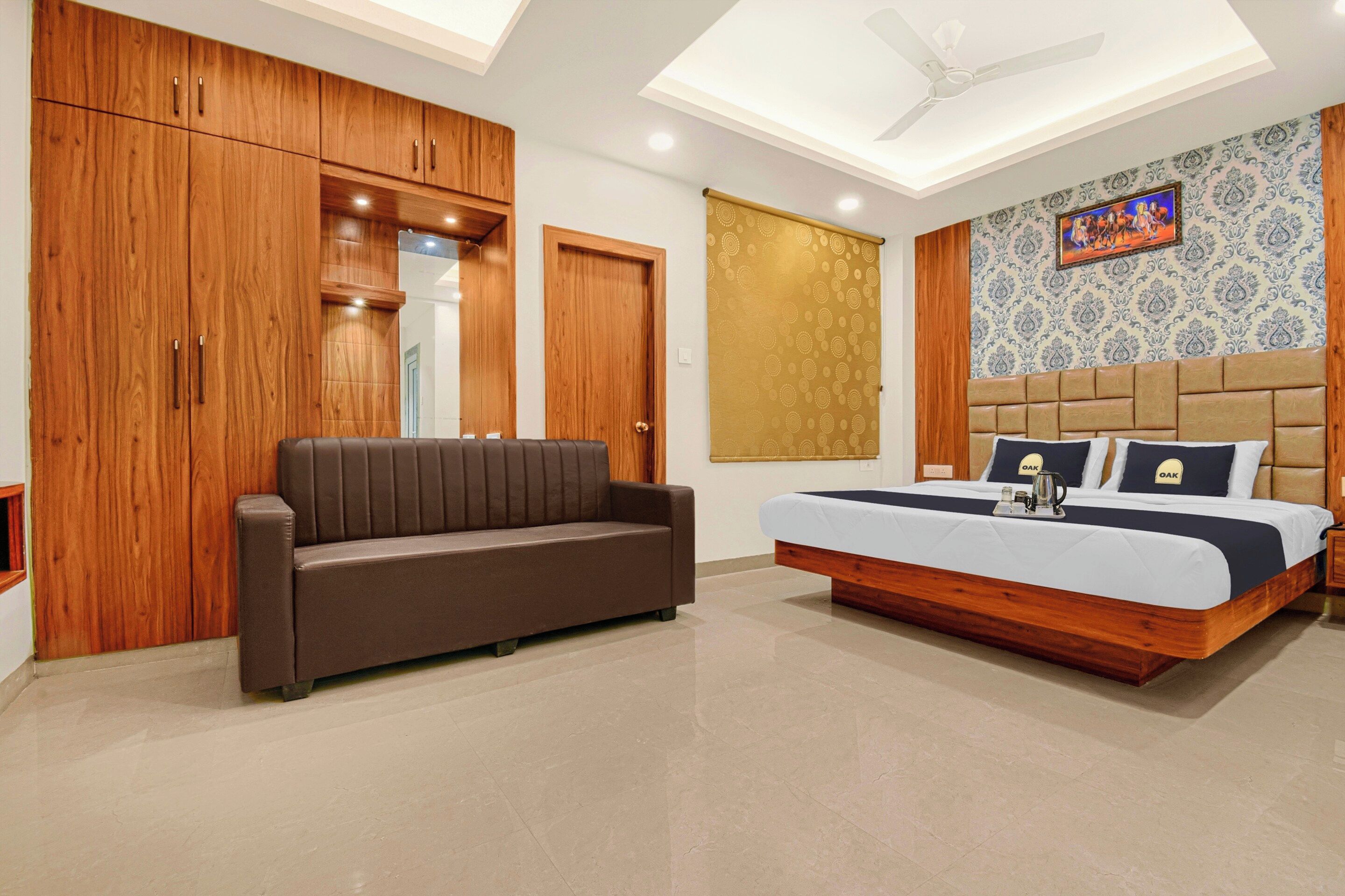 A bedroom with a bed, a sofa, wooden wardrobes, a ceiling fan, and a patterned wall.