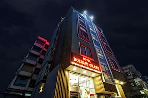 Un immeuble hôtelier à plusieurs étages, doté d’une enseigne lumineuse indiquant « HÔTEL HOLIDAY GRAND » et « CHAMBRES-RESTAURANT-BAR ».