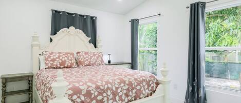 A bedroom with a bed, a nightstand, a window with curtains, and a view of greenery outside.