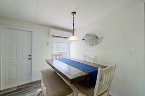 Uma sala de jantar com mesa de madeira, cadeiras, luminária pendente, aparelho de ar condicionado, janela com persianas e uma porta.