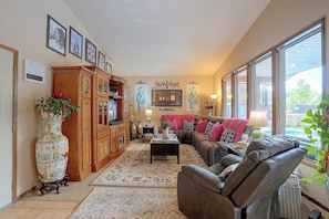 A well-lit living room with a sofa, armchairs, a coffee table, and a cabinet. There are framed pictures on the wall, a large window, and a door.