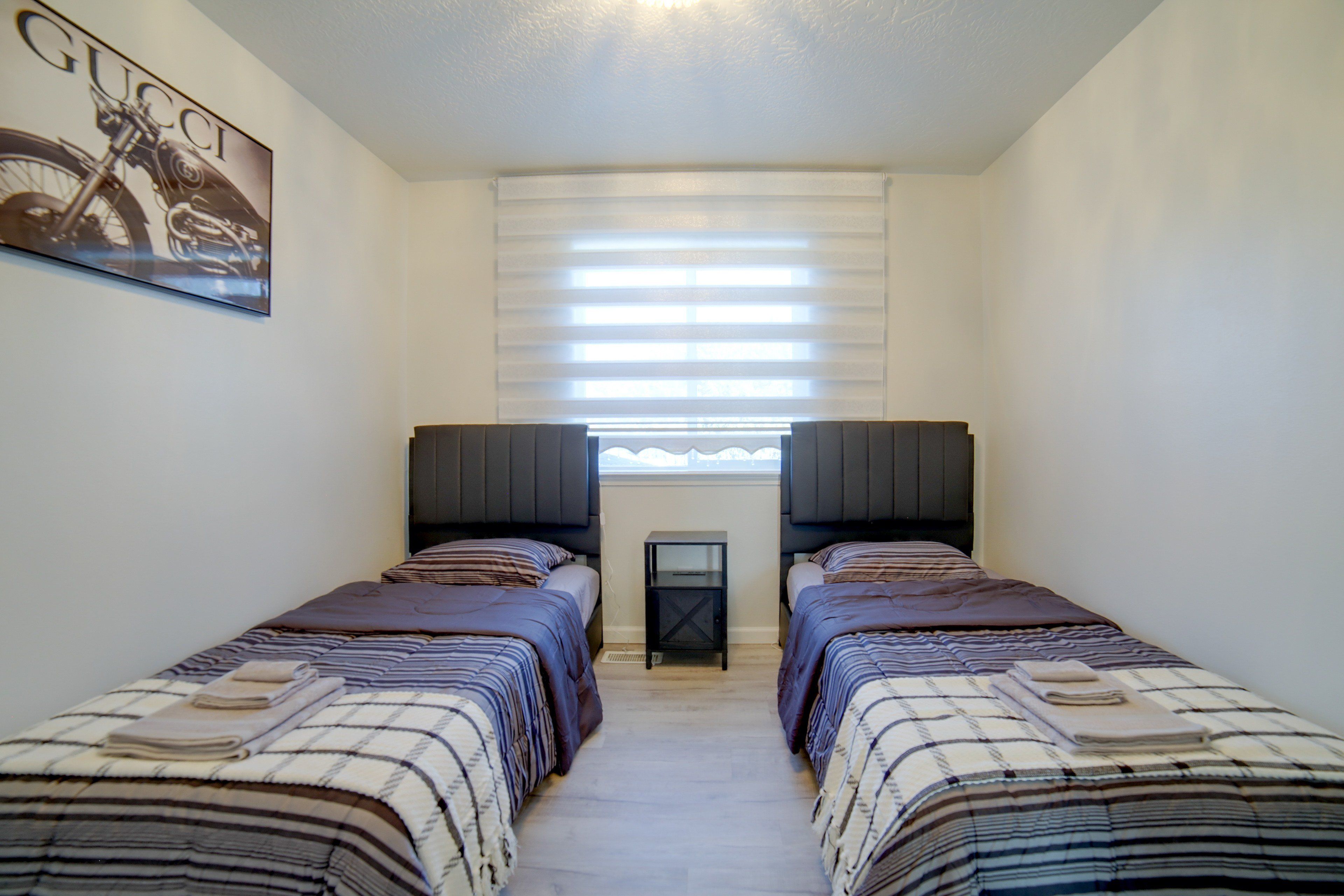 A bedroom with two beds, a window with blinds, a small table, and a wall-mounted picture.