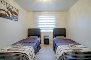 Un dormitorio con dos camas, una ventana con persianas, una mesita y un cuadro colgado en la pared.