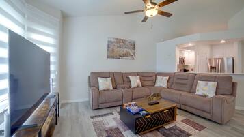 A modern living room with a sectional sofa, a flat-screen TV, a ceiling fan, and a kitchen in the background.