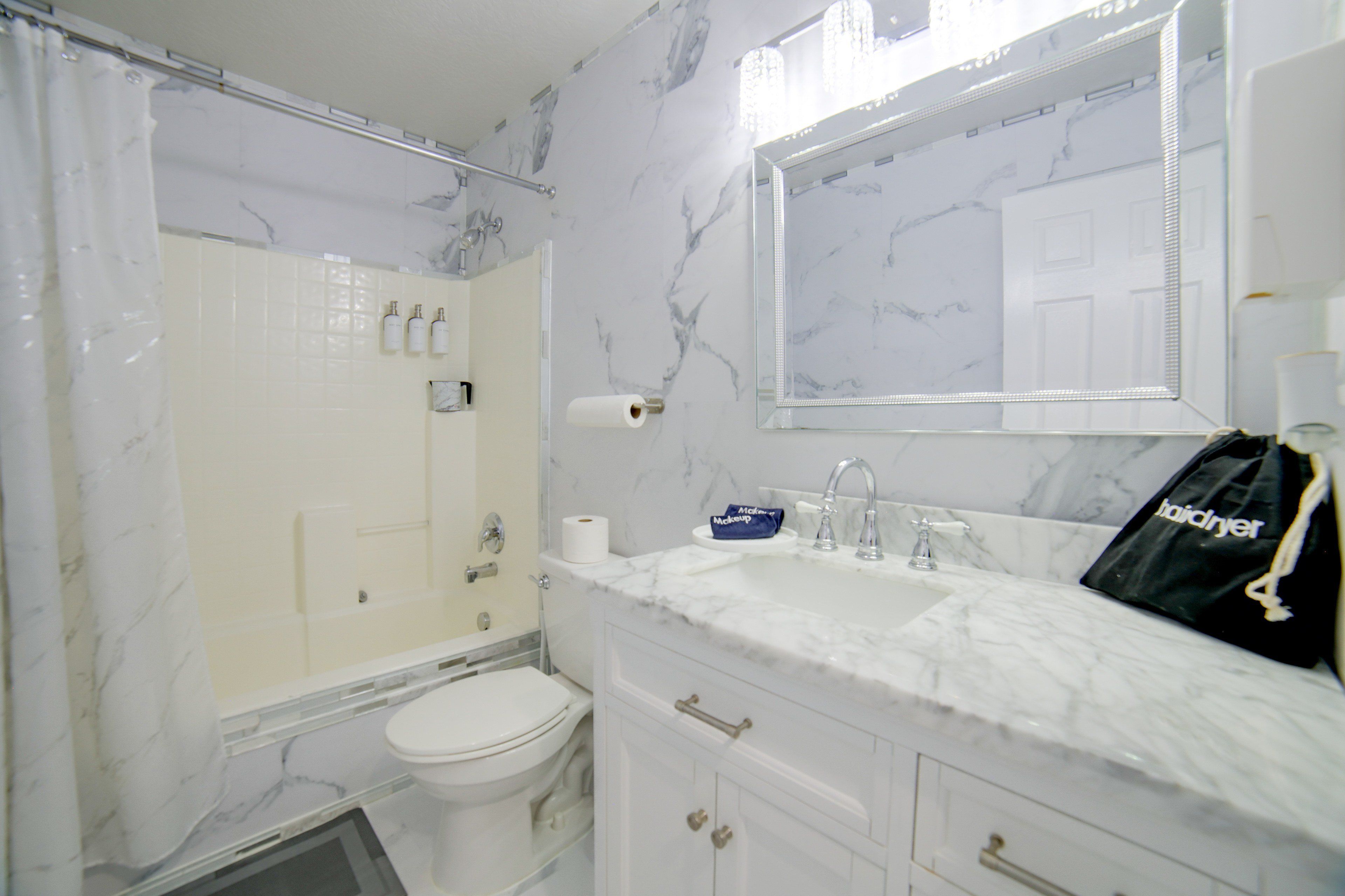 A bathroom with a white toilet, a shower with a glass door, a sink with a mirror above it, and a bag hanging on the wall.