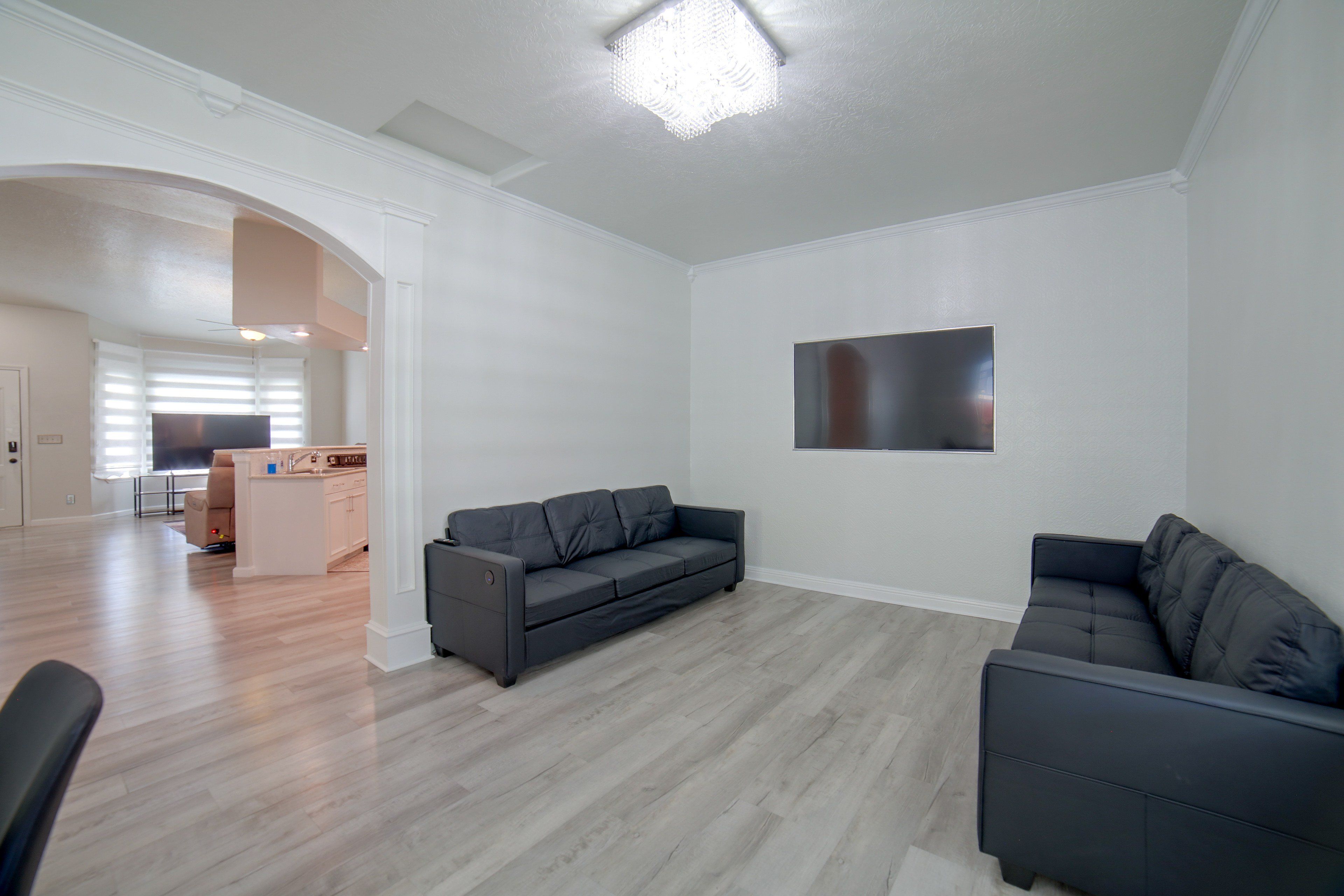A modern living room with a grey sofa, a flat-screen TV mounted on the wall, and a kitchen area in the background.