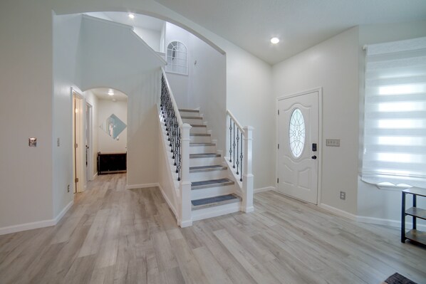 Un pasillo amplio con piso de madera, una escalera blanca con barandales negros y una puerta con un panel de vidrio decorativo.