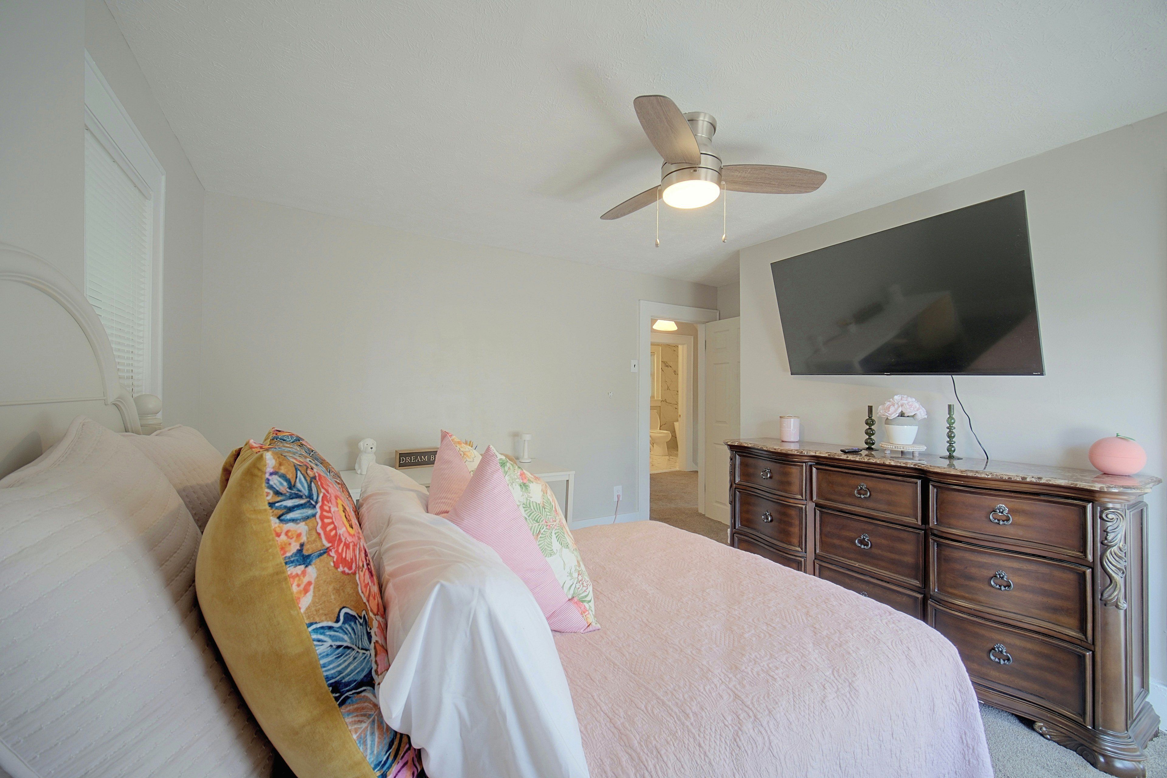 A bedroom with a bed, dresser, ceiling fan, and a TV on the wall.