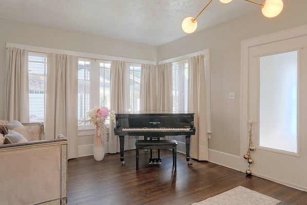 A spacious room with a grand piano, large windows with curtains, a white door, and a wooden floor. - 1 Mi to Akron Zoo! Charming Home w/ Yard (Akron)