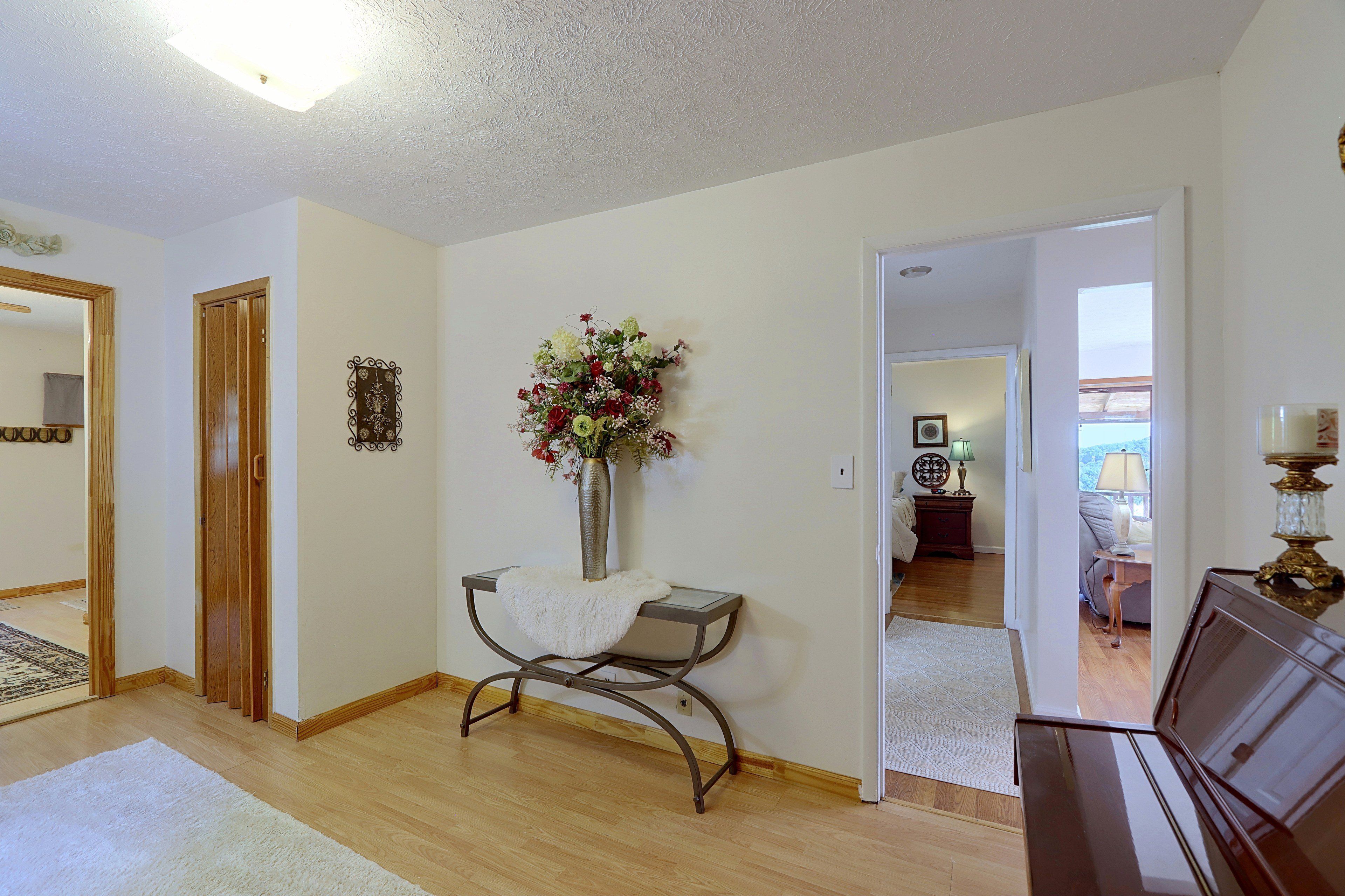A room with a wooden floor, a white wall, a door, a mirror, a vase with flowers, a table with a white cloth, and a lamp.