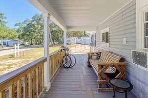 Un porche cubierto con un banco de madera y una bicicleta, con vistas a un jardín con árboles y una casa a lo lejos.