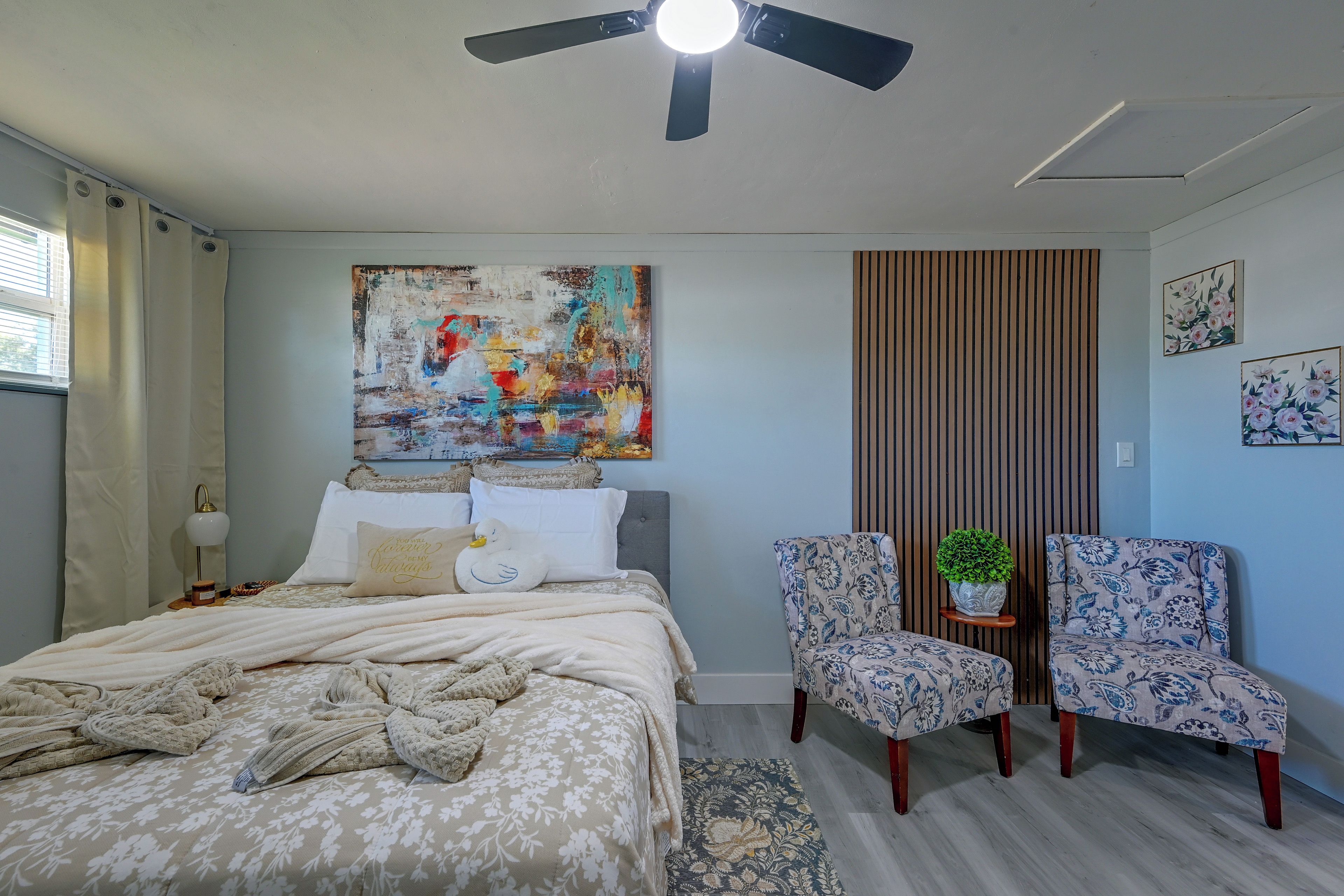 A bedroom with a bed, two chairs, a ceiling fan, a painting, and a window with curtains.