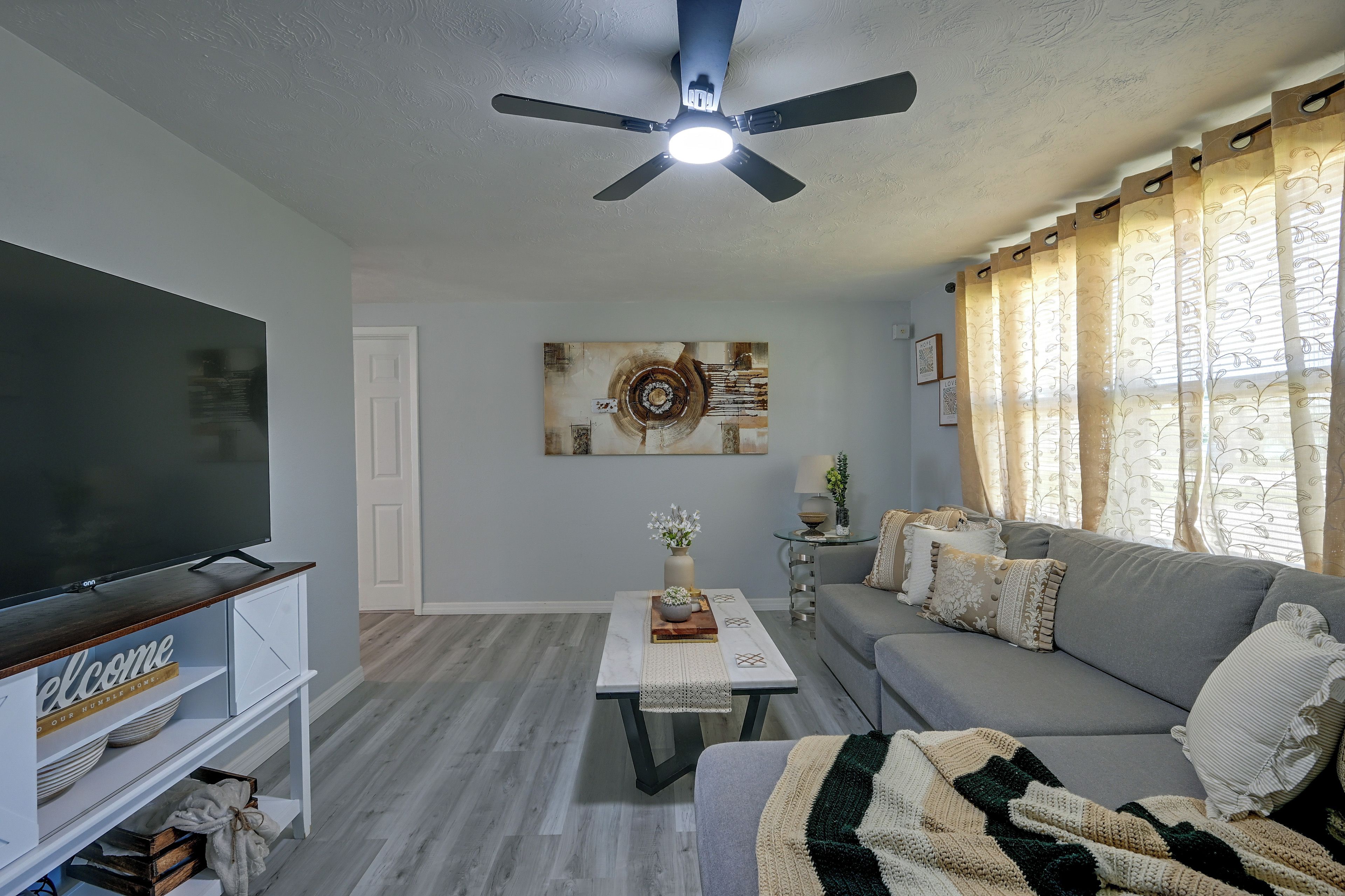 A living room with a flat-screen TV, a ceiling fan, a sofa, a coffee table, and a decorative wall piece.