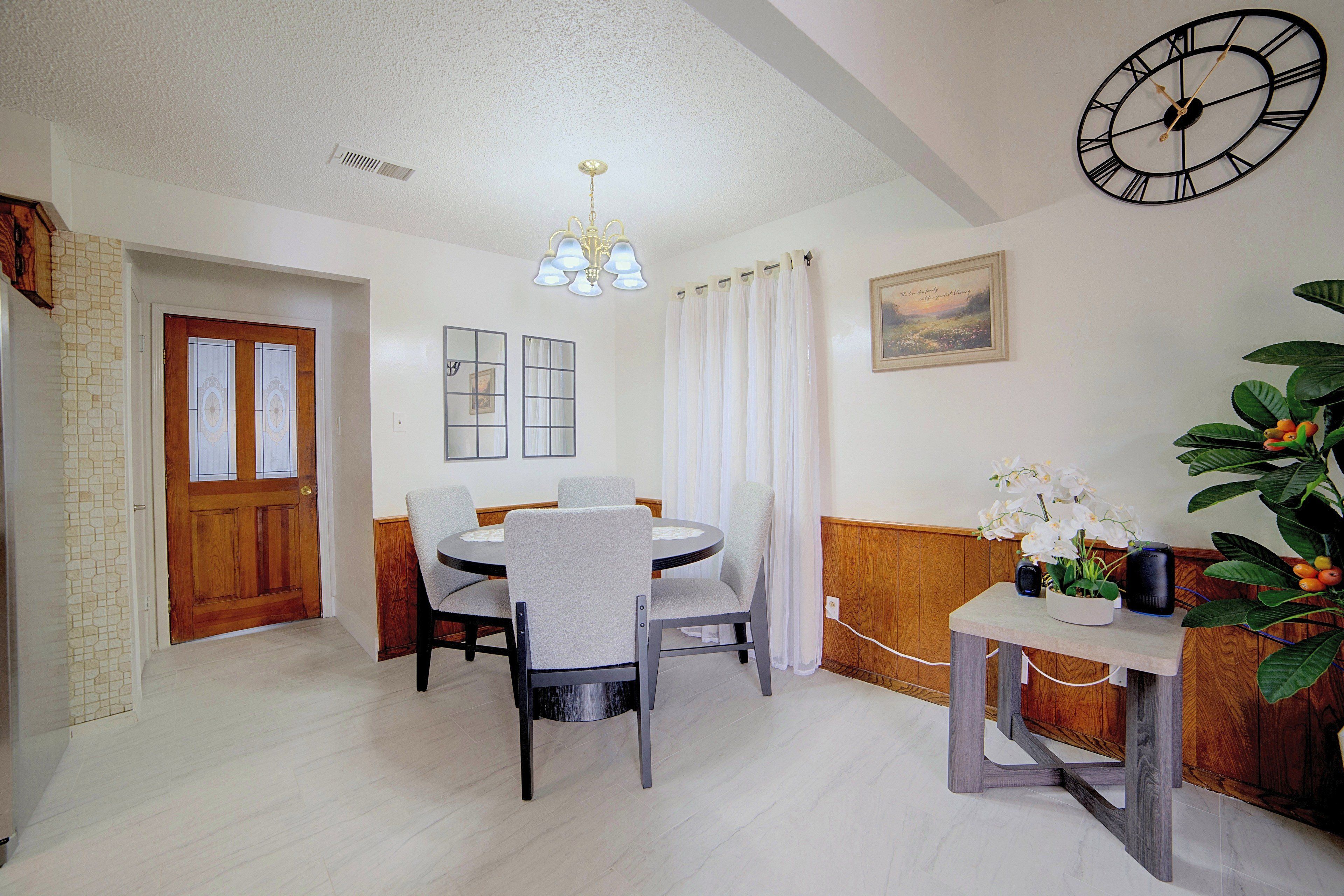 A dining area with a round table, chairs, a chandelier, a clock, and a plant.