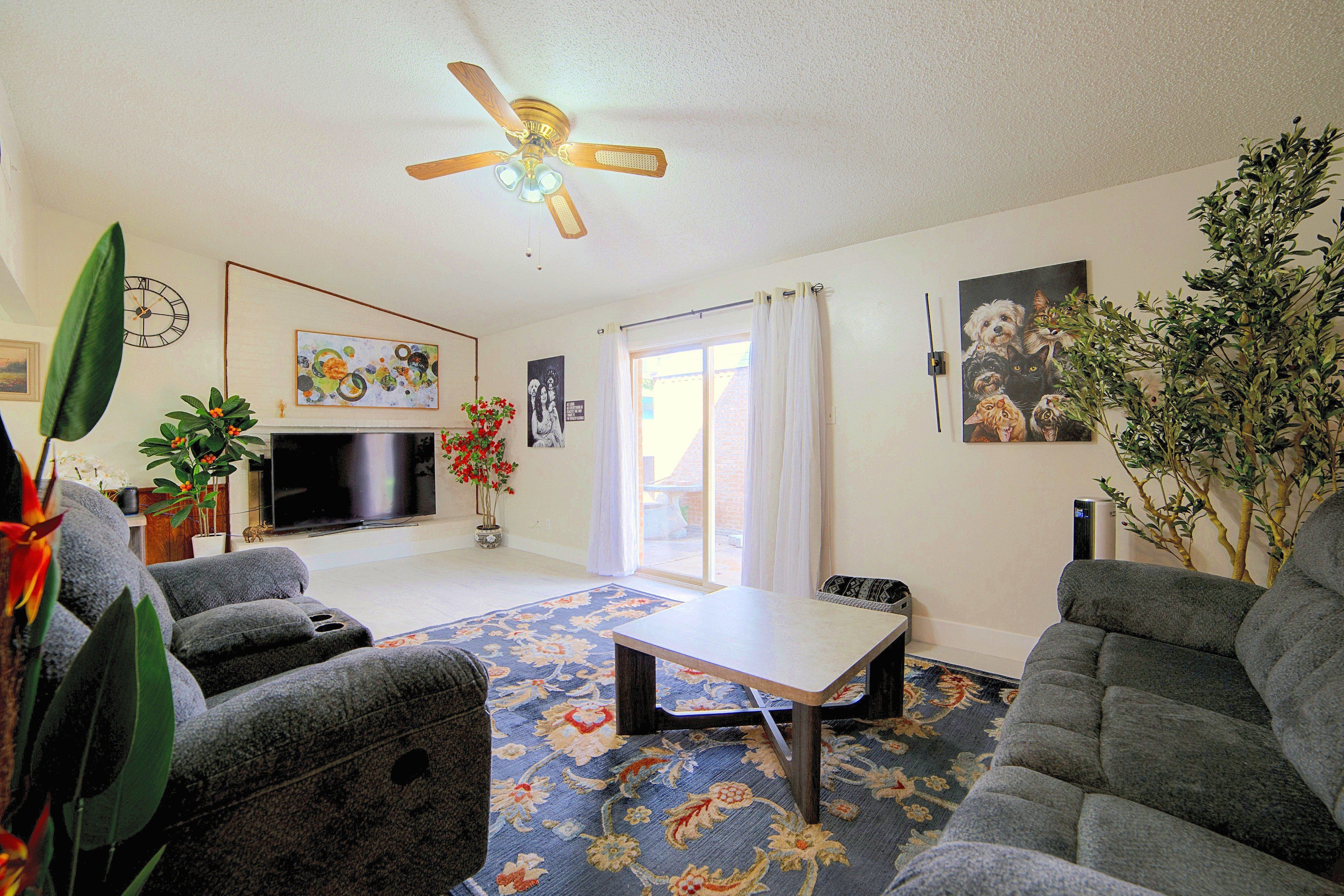 A living room with a ceiling fan, a flat-screen TV, a coffee table, two sofas, a patterned rug, and various wall decorations including paintings and a clock.