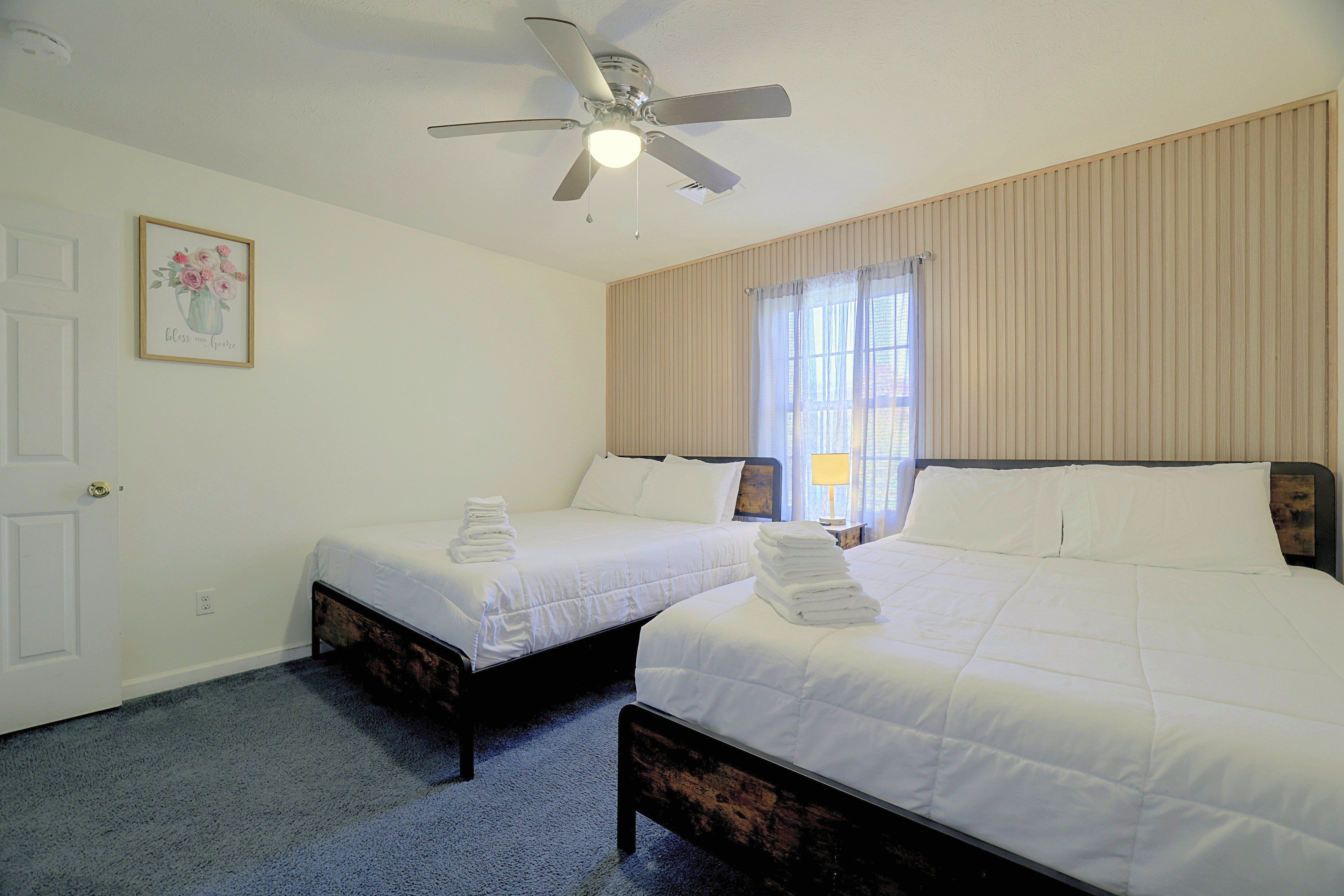 Quarto de hotel com duas camas, um ventilador de teto, uma moldura com imagem na parede e uma janela com cortinas.