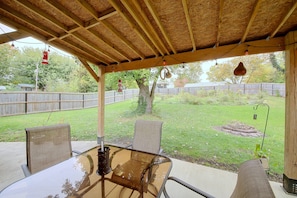 Un patio exterior con una mesa y sillas de vidrio, techo de madera y vista a un jardín trasero con cercado de madera y árboles.