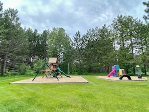 A playground with a wooden playhouse and slides, surrounded by a grassy area and trees. - Trout Creek 13 | Pools, Hot Tub, Great Location! (Harbor Springs)