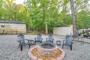 A gravel patio with a fire pit surrounded by Adirondack chairs, a wooden cabin, and a shed in a wooded area.