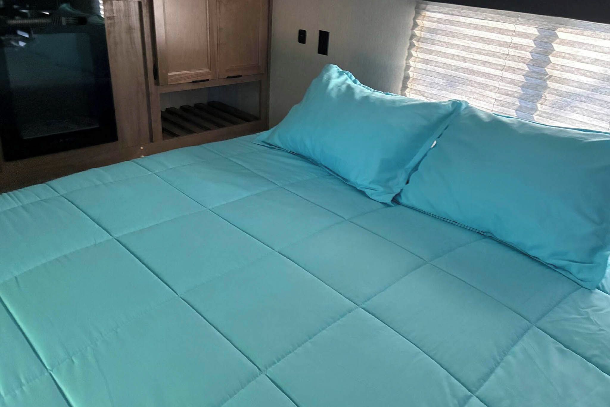 Un lit avec une couette et des oreillers bleu clair, des armoires en bois et une fenêtre avec des stores.