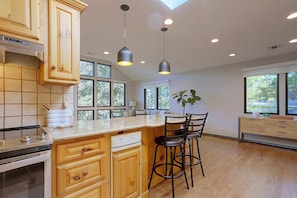 A modern kitchen with wooden cabinetry, a central island with bar stools, pendant lighting, and large windows offering views of the outdoors.