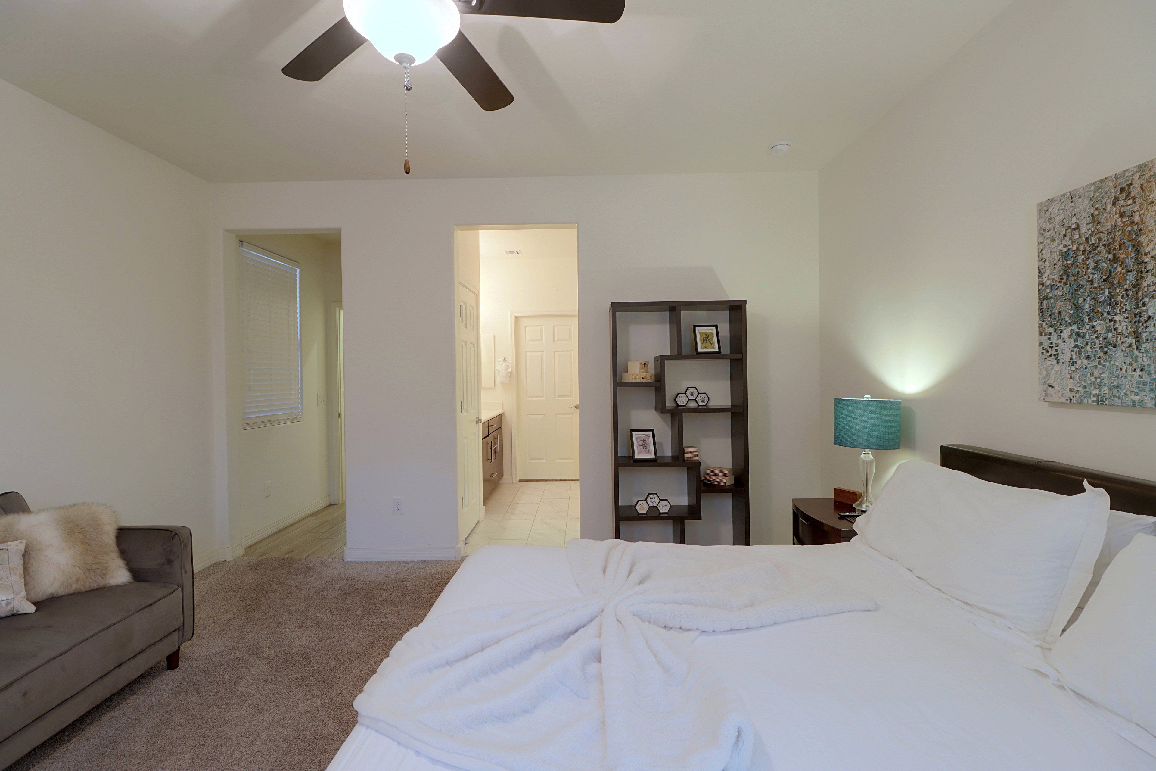 Quarto de hotel com cama, mesa de cabeceira, luminária, estante de livros e ventilador de teto.