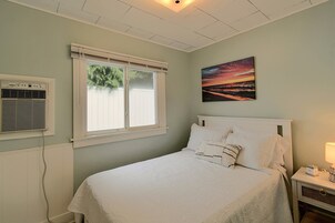 Un dormitorio con cama, ventana, equipo de aire acondicionado, mesita de noche y un cuadro colgado en la pared.