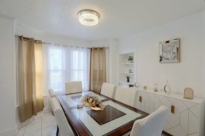 A dining area with a glass-top table, white chairs, a chandelier, and a view of the outdoors through the window.