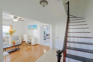 A spacious living area with a wooden floor, a white sofa, a desk, and a staircase with a dark wooden handrail.