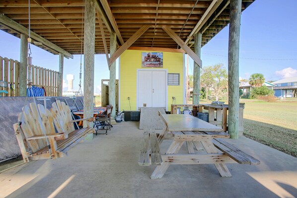 A covered outdoor area with a wooden table and benches, a door, and a sign advertising a house for sale.
