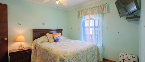 A bedroom with a bed, a nightstand, a lamp, a ceiling fan, a television, and a window with curtains.