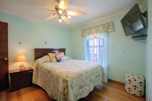 A bedroom with a bed, a nightstand, a lamp, a ceiling fan, a television, and a window with curtains.