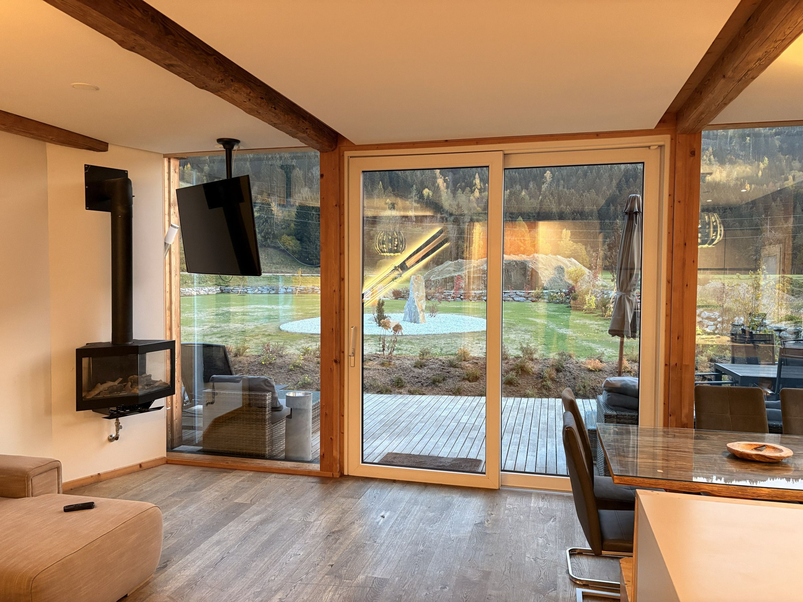 Una sala de estar moderna con chimenea, televisor y puerta corredera de vidrio que da a un patio exterior con terraza de madera y vistas a un área boscosa.