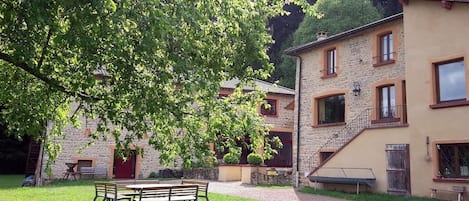 Un patio con un banco de madera, edificios de piedra y un árbol que da sombra.