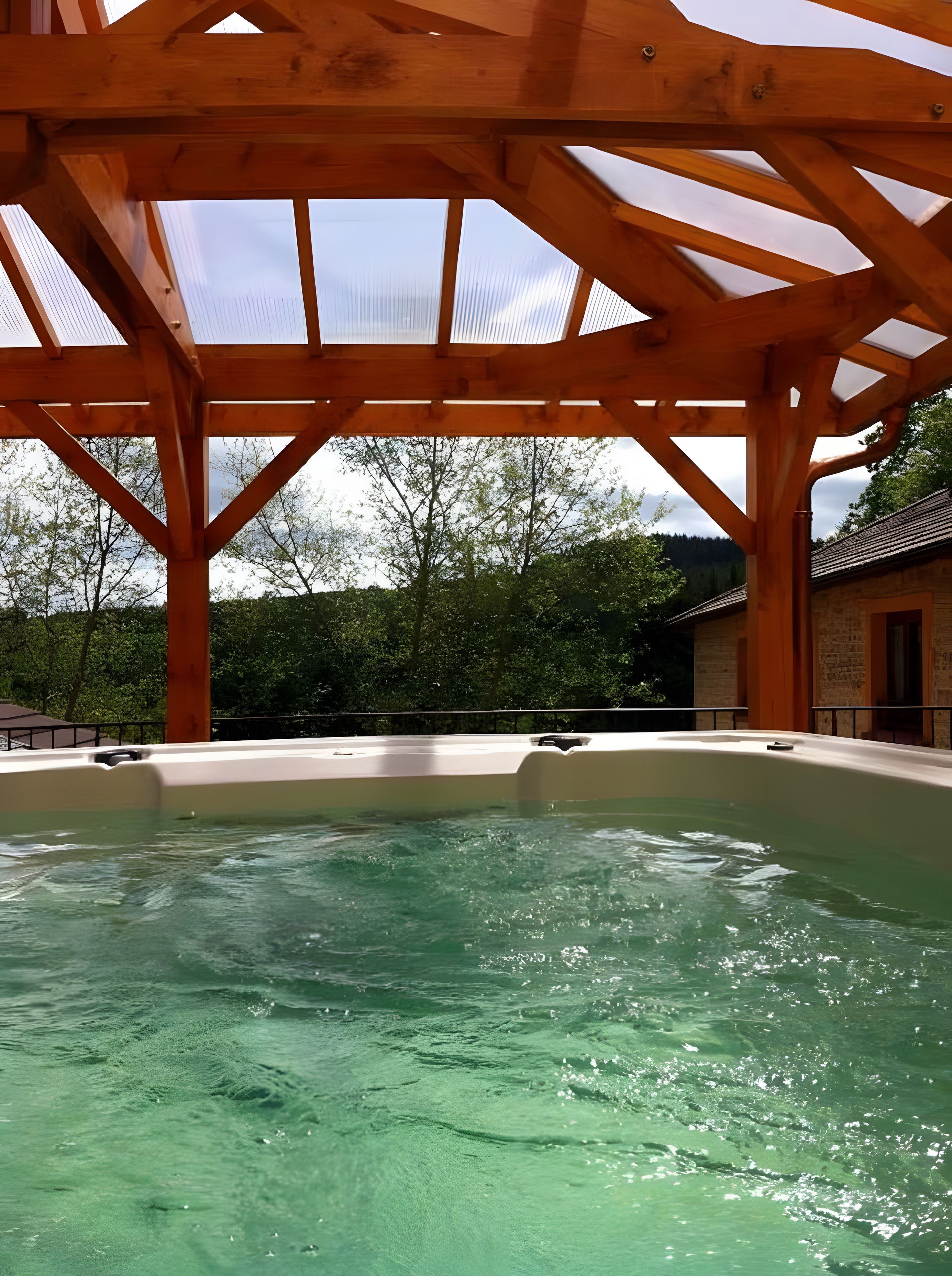 Een hottub onder een houten pergola, met uitzicht op bomen en een gebouw op de achtergrond.