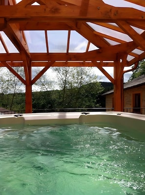 Een hottub onder een houten pergola, met uitzicht op bomen en een gebouw op de achtergrond.