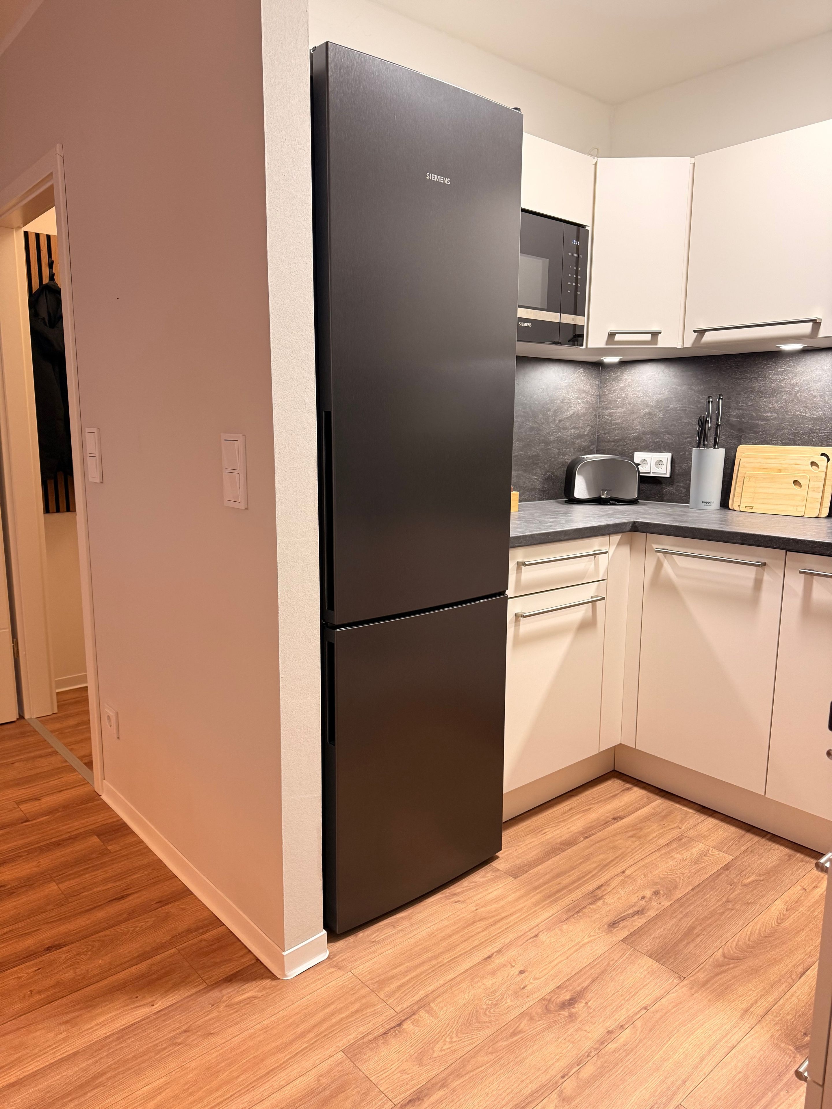 Eine Küche mit einem schwarzen Kühlschrank, weißen Schränken, einer Mikrowelle und einem Holzer Schneidebrett.