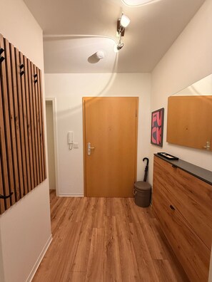 Un pasillo con una puerta de madera, un perchero, un espejo y un cubo de basura pequeño.
