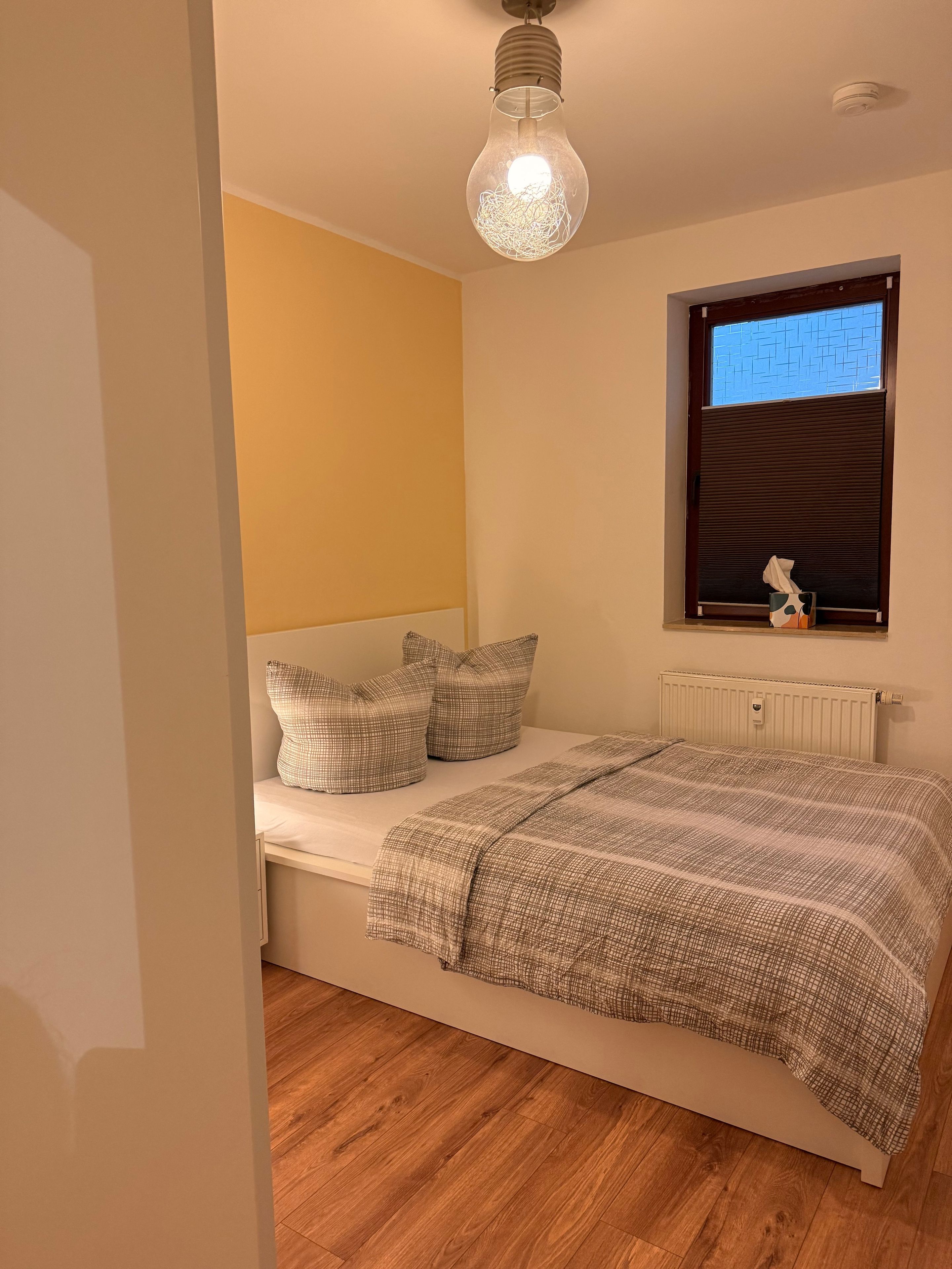 Ein Schlafzimmer mit einem Bett, einem Heizkörper, einem Fenster mit Jalousien und einer Pendelleuchte.
