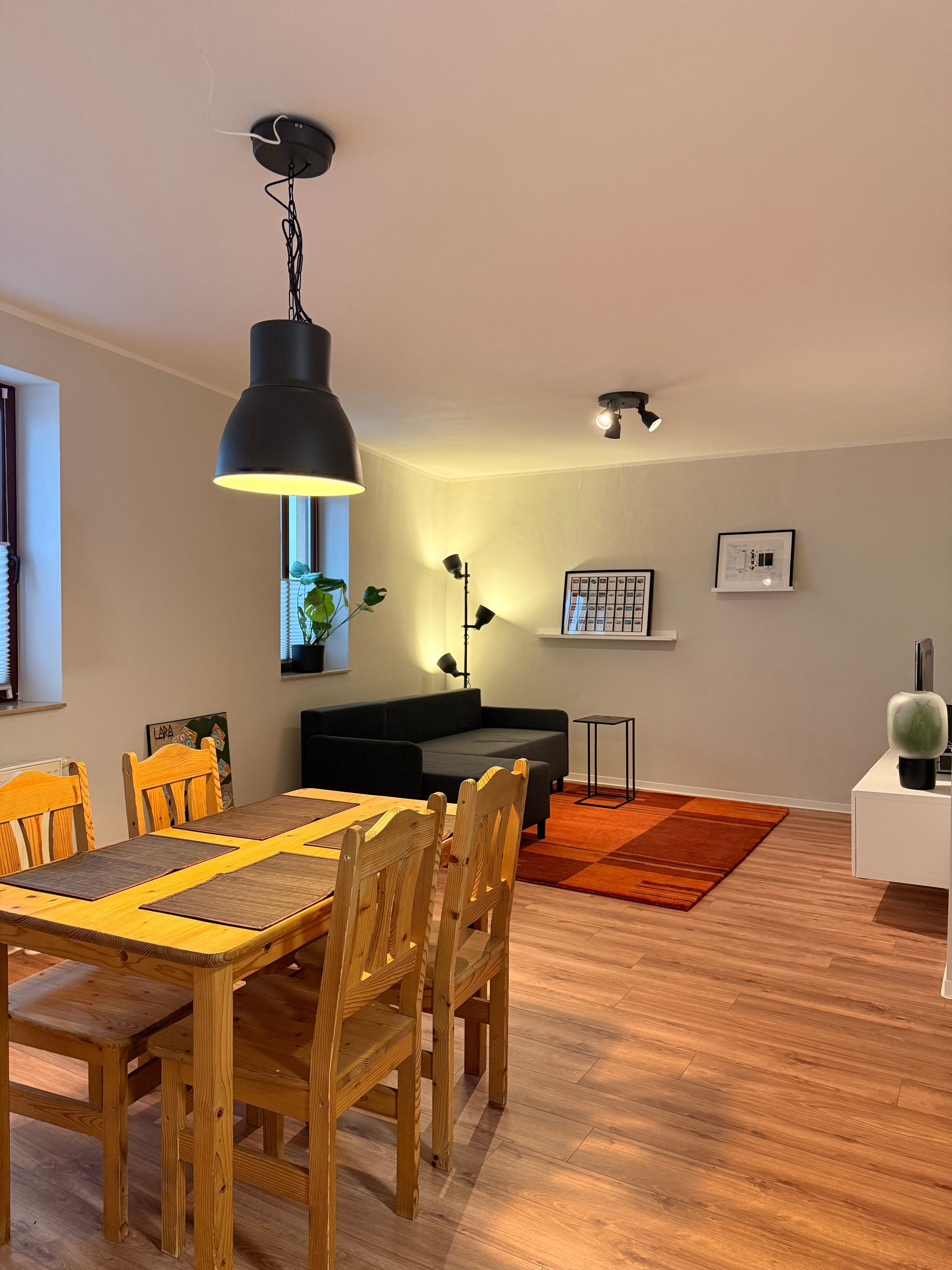 Ein Essbereich mit einem Holztisch, Stühlen, einer schwarzen Pendelleuchte und einem schwarzen Sofa im Hintergrund.