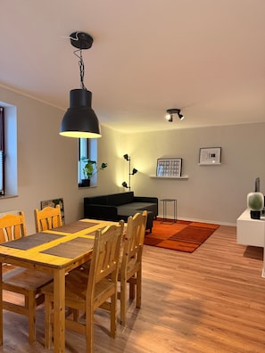 Un área de comedor con una mesa y sillas de madera, una lámpara colgante negra y un sofá negro al fondo.