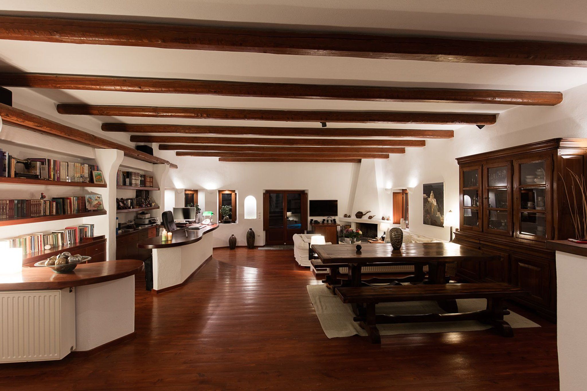 Une pièce spacieuse avec des poutres en bois au plafond, une longue table à manger et un coin cuisine doté de meubles en bois et d’une bibliothèque.
