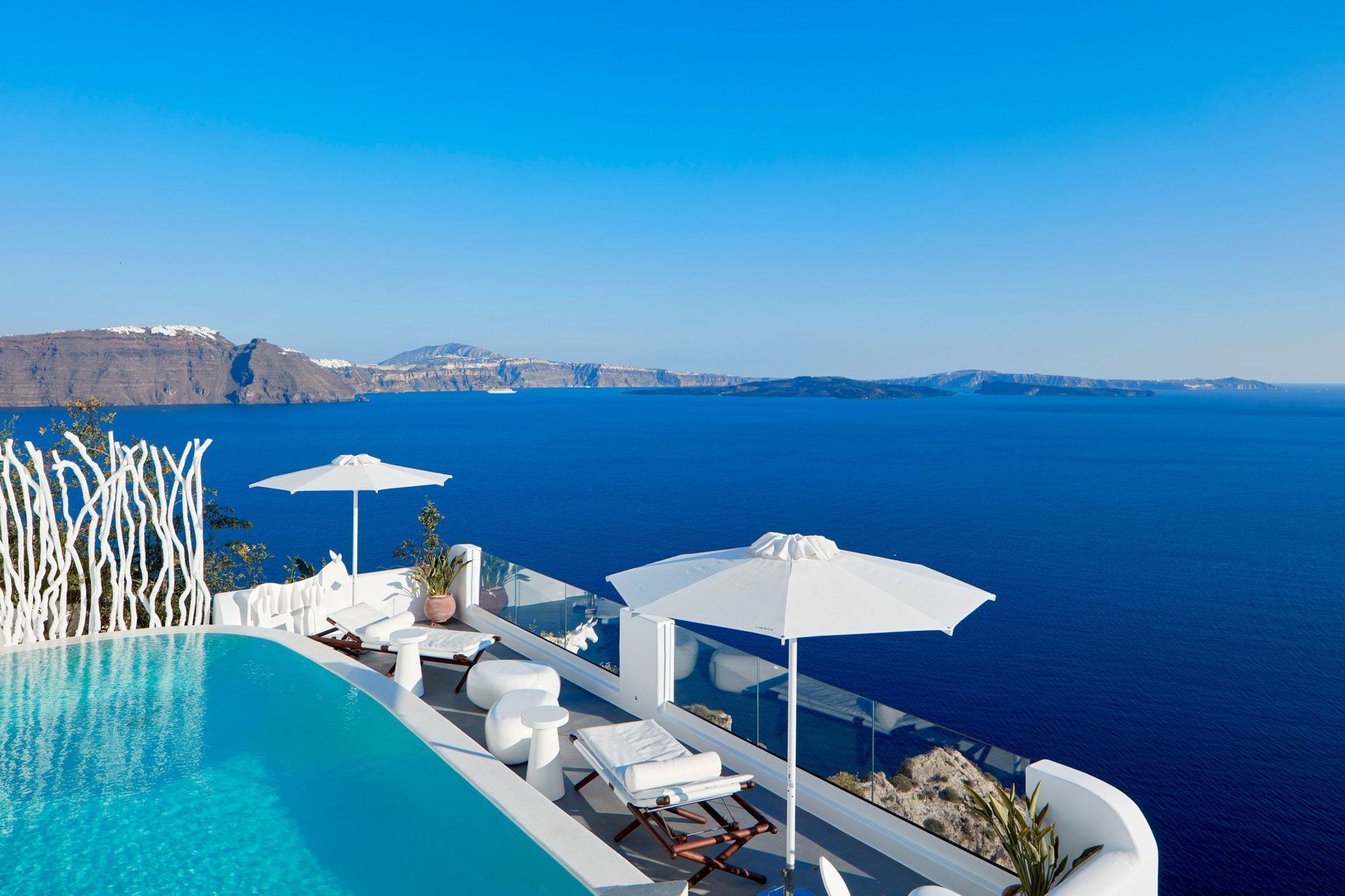 Un espace aménagé au bord de la piscine, avec des chaises longues et des parasols, donnant sur une étendue d’eau et offrant une vue sur une côte lointaine.
