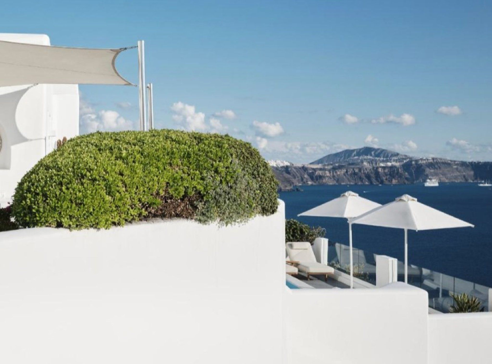 Un bâtiment blanc surmonté d’un jardin-terrasse verdoyant, avec des parasols blancs, et offrant une vue sur la mer et les montagnes.