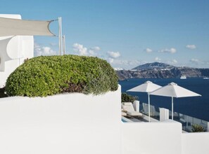 Un bâtiment blanc surmonté d’un jardin-terrasse verdoyant, avec des parasols blancs, et offrant une vue sur la mer et les montagnes.