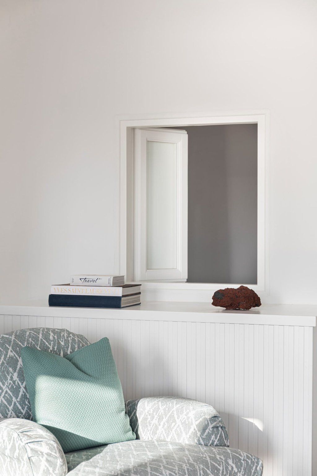 Une pièce avec un mur blanc, une fenêtre à cadre blanc, un radiateur, une pile de livres et un coussin sur une chaise.